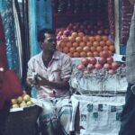 man in white and gray robe sitting on white plastic crate with orange fruits